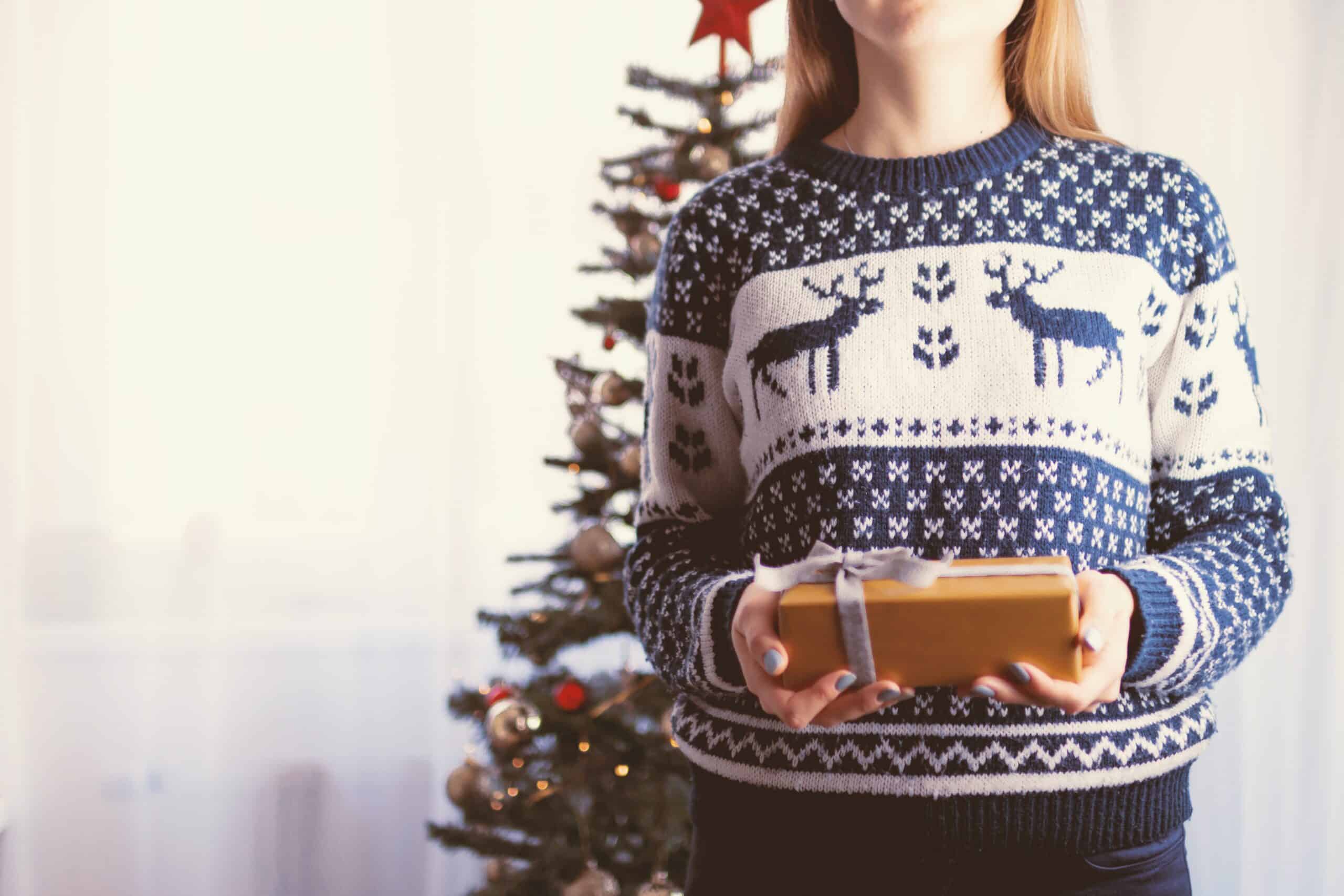 woman in christmas sweater holding wrapped gift in front of christmas tree