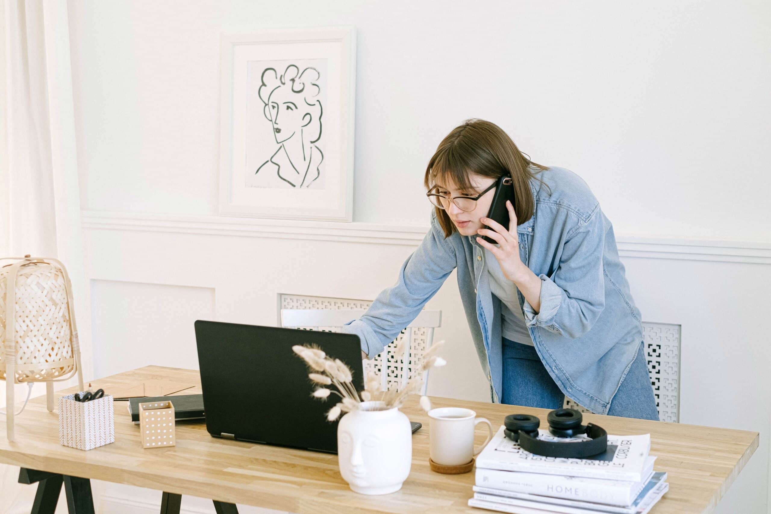 woman standing with a phone to her ear and looking at her open laptop on her desk