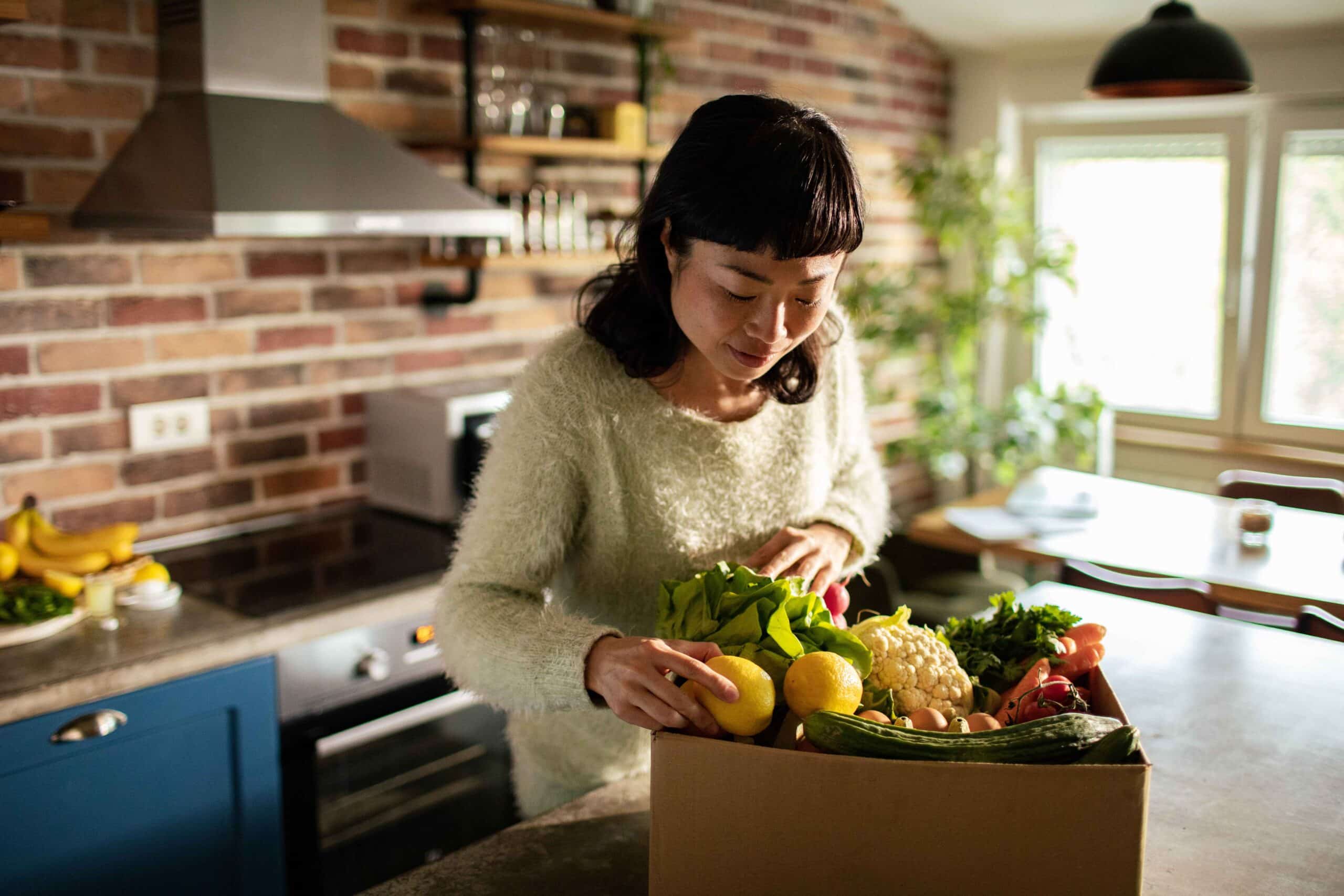 Household assistant taking vegetables out of cardboard box in kitchen