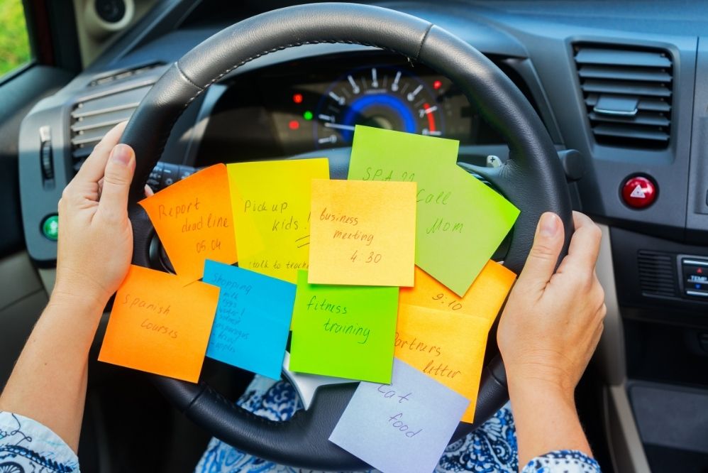 Overwhelmed parent driving with sticky notes on steering wheel.